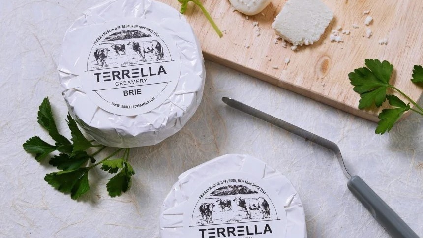 Dva kulaté sýry brie značky Terella Creamery leží na stole. Jeden z nich je na dřevěném prkénku vedle nože na sýr a čerstvé petrželky.