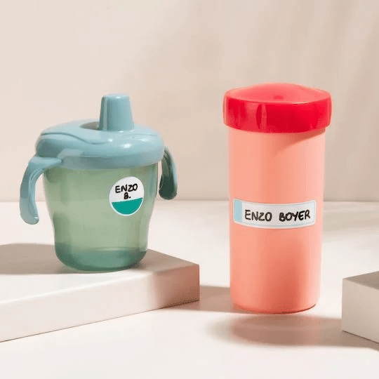 Zelený průhledný plastový kelímek s hubičkou a držadly a lososově zbarvená podlouhlá plastová láhev s červeným uzávěrem, na které jsou nalepeny samolepky s rukopisem.