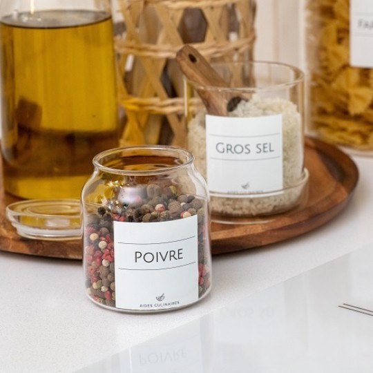 Dvě skleněné nádoby s bílými štítky, jedna na bílé desce stolu, druhá na dřevěném stojanu s rustikálními prvky v pozadí.