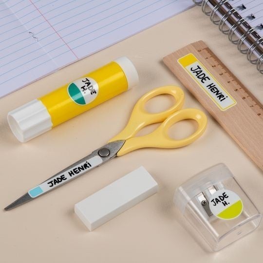 Dřevěný stůl, na kterém leží lepidlo, nůžky, pravítko a plastová krabička s pery, na kterých jsou nálepky na psaní rukou, a vedle ní bílá guma.