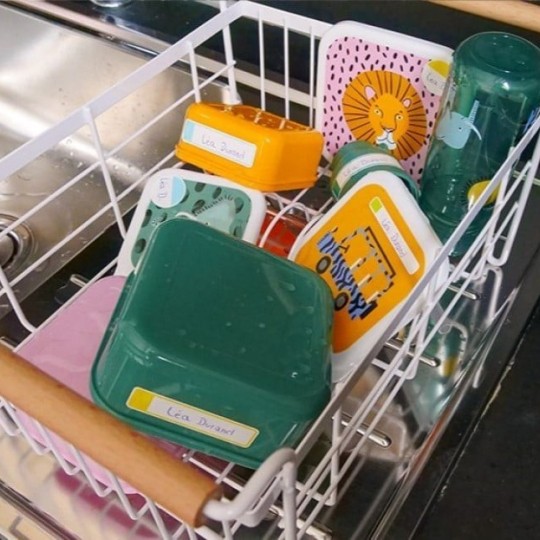 Kovový koš zavěšený nad dřezem, ve kterém leží několik různých plastových snídaňových misek a bidonů s dětskými motivy.