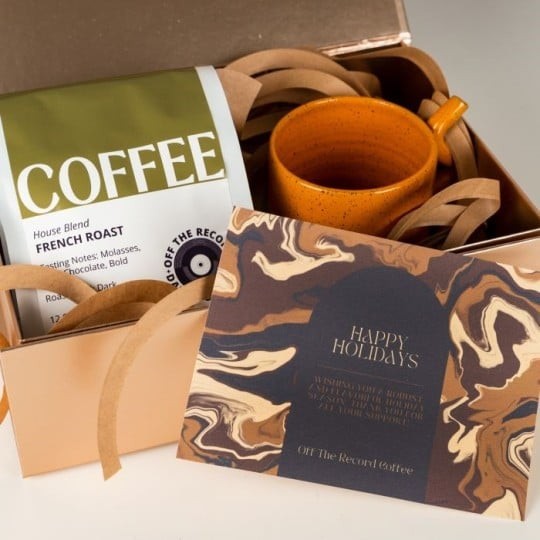 Dárková krabička s balením kávy French Roast, oranžovým keramickým hrnkem a přiloženou sváteční kartičkou s nápisem „Happy Holidays“ na mramorovaném pozadí.