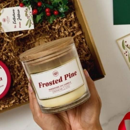 Ruce držící svíčku „Frosted Pine“ nad otevřenou dárkovou krabičkou s dalším svátečním zbožím a dekorací s cesmínou.