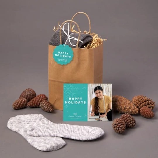 Dárková papírová taška s etiketou „Happy Holidays“, šedé ponožky, přání s fotografií ženy a nápisem „Happy Holidays“, vše obklopené šiškami na šedém pozadí.