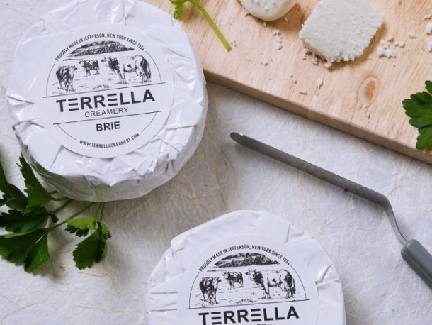Dva kulaté sýry brie značky Terella Creamery leží na stole. Jeden z nich je na dřevěném prkénku vedle nože na sýr a čerstvé petrželky.