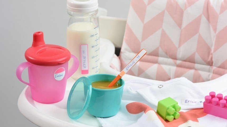 Růžová plastová láhev s červenou hubičkou, zelený pastelový kelímek, plastová láhev na mléko a různé hračky a dětské potřeby na bílém plastovém stole.