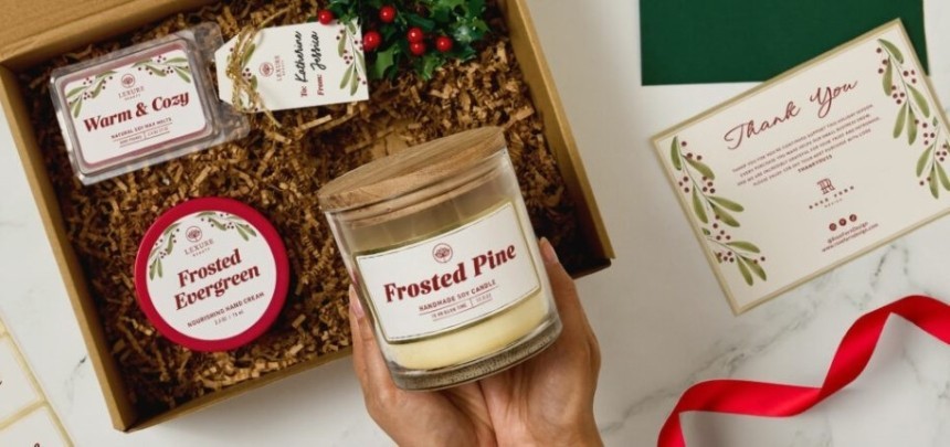 Dárková krabička se svátečními produkty na přírodním podkladu: v ruce je svíčka "Frosted Pine", v krabičce jsou krémy a svíčka s nápisy "Warm & Cozy" a "Frosted Evergreen", větvička s červenými bobulemi a děkovná kartička s ozdobnými motivy a červenou stuhou.