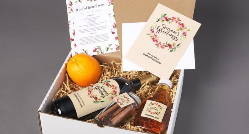 Bílá dárková krabice s lahví vína, sklenicí se skořicí, lahvičkou medu, pomerančem a sváteční recept a kartička s věncem a nápisem „Season’s Greetings“.