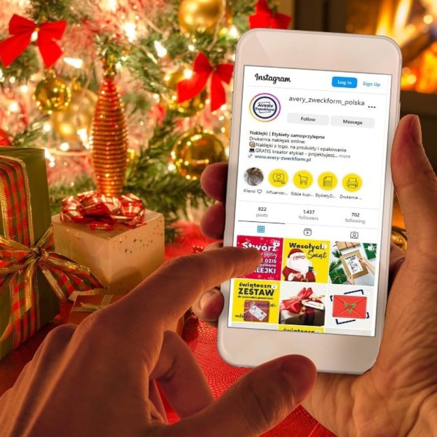 Osoba držící smartphone s otevřeným Instagramem před vánočně ozdobeným stromkem a dárky zabalenými v barevném papíru a mašlích.