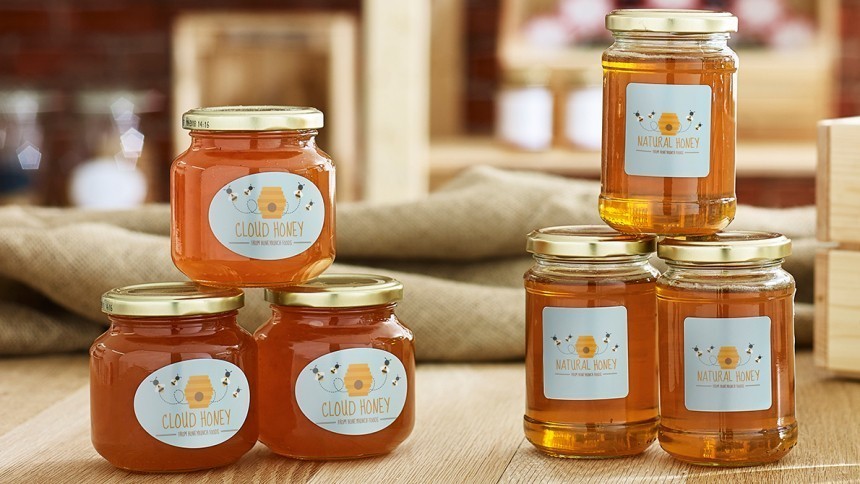 Několik skleněných sklenic s světle hnědým medem, postavených na dřevěné desce; na každé sklenici je etiketa s nápisem „Cloud Honey” nebo „Natural Honey” a obrázkem úlu.