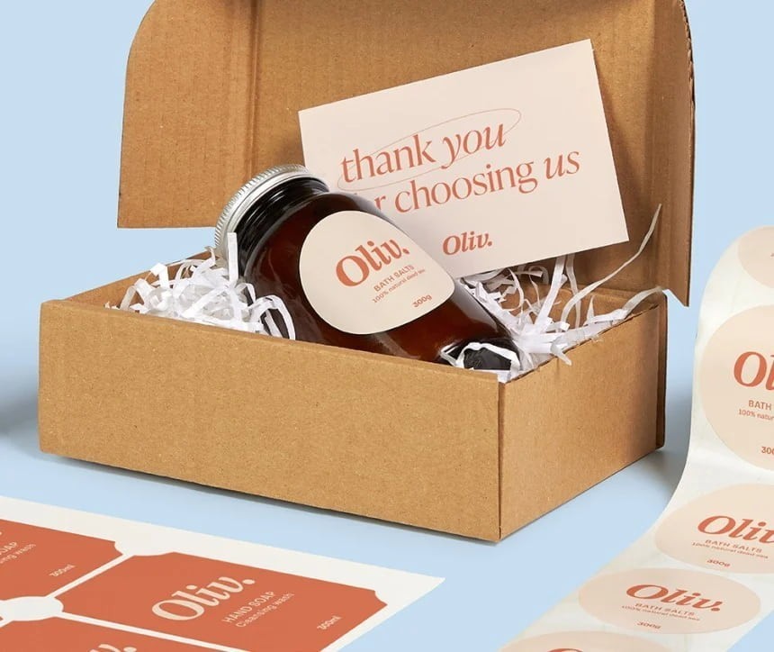 Otevřená kartonová krabice s výplní z bílých papírových proužků, ve které leží skleněná dóza s etiketou „Oliv.” a kartička s poděkováním „thank you for choosing us“. Vedle krabice jsou vidět archy a role kulatých etiket se stejným logem.