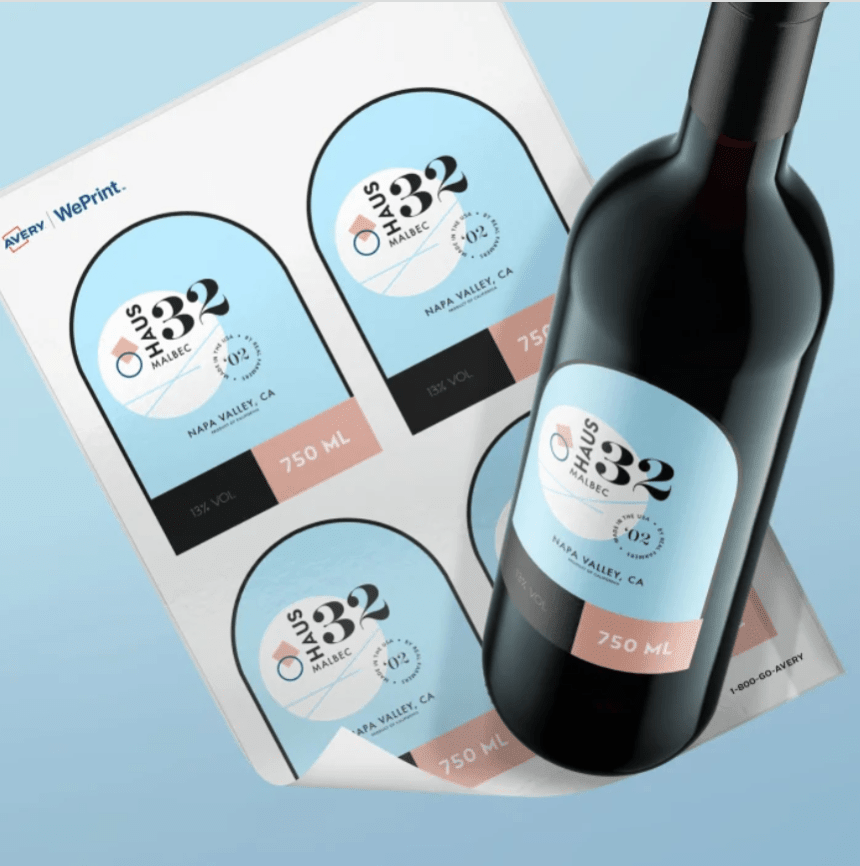 Láhev červeného vína s modrou etiketou „Haus 32 Malbec“ stojí na archu samolepicích etiket, na kterém jsou viditelné čtyři stejné, dosud nepoužité etikety.