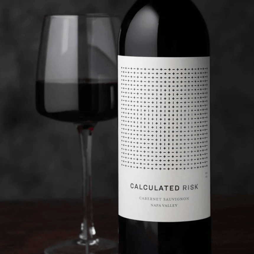 Detail láhve červeného vína s minimalistickou bílou etiketou „CALCULATED RISK – CABERNET SAUVIGNON – NAPA VALLEY“, v pozadí stojí naplněná sklenka vína.