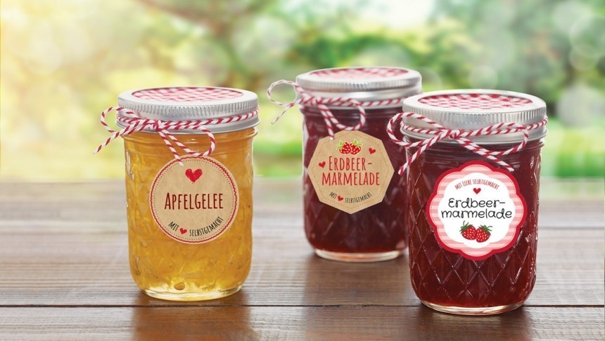 Tři skleněné zavařovací sklenice s domácími výrobky – jedna se světlým jablečným želé a dvě s červeným jahodovým džemem – stojí na dřevěné desce. Každá sklenice má kovové víčko, ozdobný provázek a barevnou etiketu s německým popisem obsahu.