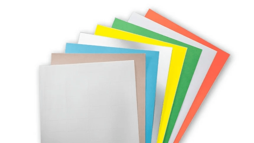 Vějíř barevných archů etiket: nahoře bílý arch s jemně vyznačenými poli na etikety a pod ním postupně béžový, modrý, žlutý, zelený, bílý a oranžový karton.