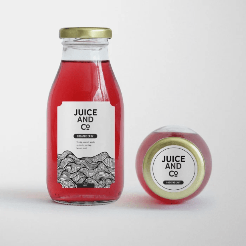 Dvě skleněné lahve s červeným džusem na bílém pozadí: jedna stojí s obdélníkovou etiketou s nápisem „JUICE AND CO – BREATHE EASY“ a černobílou grafikou vln, druhá leží naležato a ukazuje kulatou etiketu se stejným logem na zlatém víčku.
