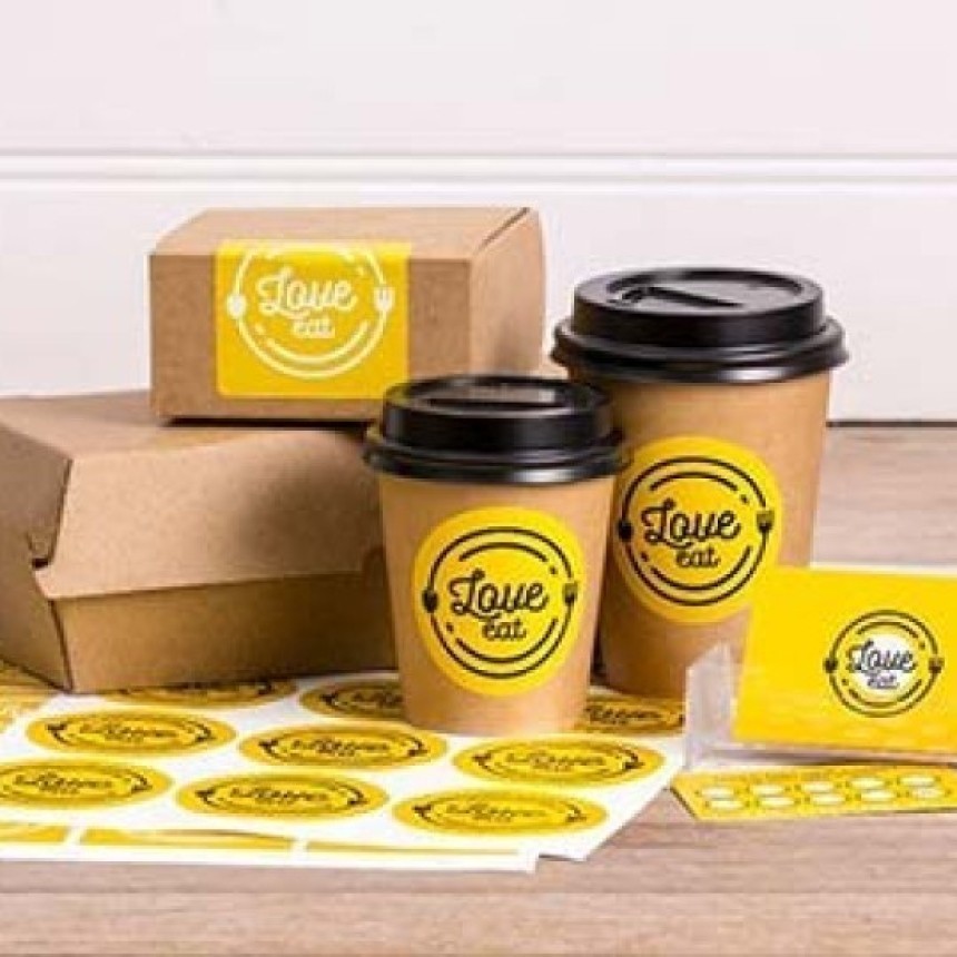 Papírové kelímky na kávu a krabičky s sebou ozdobené žlutými kulatými samolepkami s logem "Love Eat". Vedle leží arch se stejnými samolepkami.