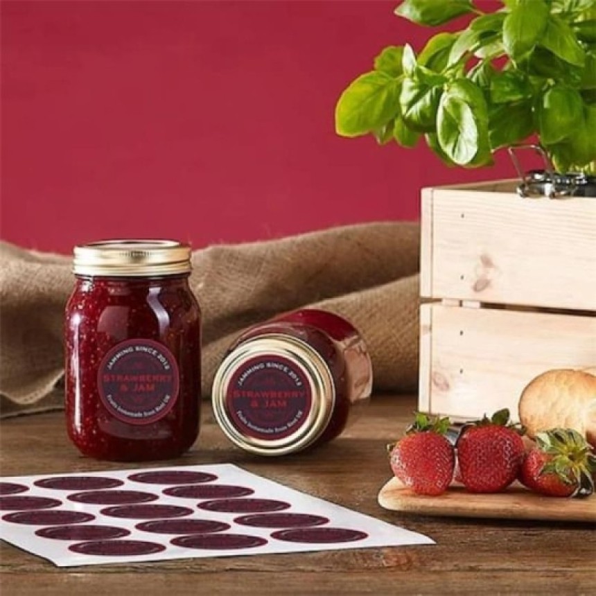 Dvě sklenice domácího jahodového džemu s etiketami "Strawberry & Jam" stojí na dřevěném stole vedle čerstvých jahod, bazalky a archu s etiketami.
