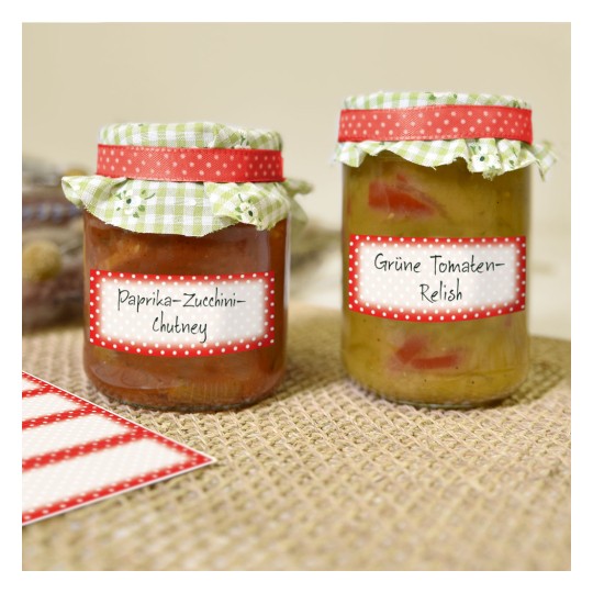 Obrázek zobrazuje dvě sklenice domácích dochucovadel. Levá sklenice je označena 'Paprika-Cuketový-Chutney' a pravá sklenice je označena 'Zelená Rajčatová-Relish.' Obě sklenice mají dekorativní víčka s kostkovaným vzorem a puntíkovanými štítky.