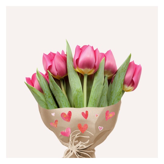 Kytice růžových tulipánů zabalená v papíře s dekorativními srdíčky, umístěná na světlém pozadí.