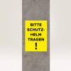 Obrázek zobrazuje žlutou ceduli na betonovém sloupu s nápisem 'BITTE SCHUTZ-HELM TRAGEN!', což se překládá jako 'Prosím, noste helmu!', což naznačuje bezpečnostní požadavek.
