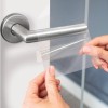 Obrázek ukazuje osobu, která nanáší průhlednou lepicí fólii na kliku dveří, která je vyrobena z nerezové oceli. Pozadí je moderní, elegantní prostředí se skleněnými dveřmi.