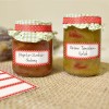 Obrázek zobrazuje dvě sklenice domácích dochucovadel. Levá sklenice je označena 'Paprika-Cuketový-Chutney' a pravá sklenice je označena 'Zelená Rajčatová-Relish.' Obě sklenice mají dekorativní víčka s kostkovaným vzorem a puntíkovanými štítky.