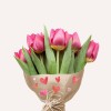 Kytice růžových tulipánů zabalená v papíře s dekorativními srdíčky, umístěná na světlém pozadí.